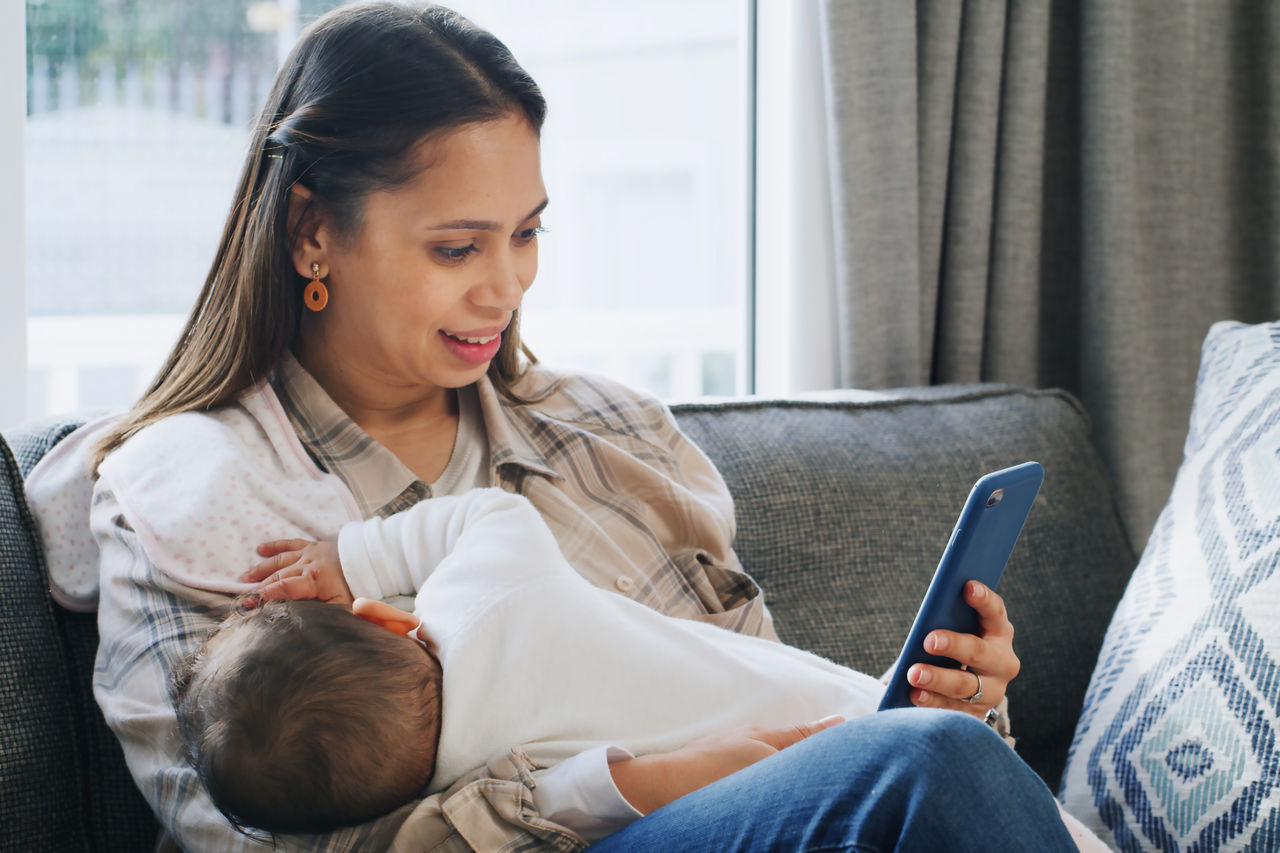 An adult sits on a couch, breastfeeding an infant while reading on a smartphone. The scene captures a moment of modern caregiving and multitasking in a home setting.