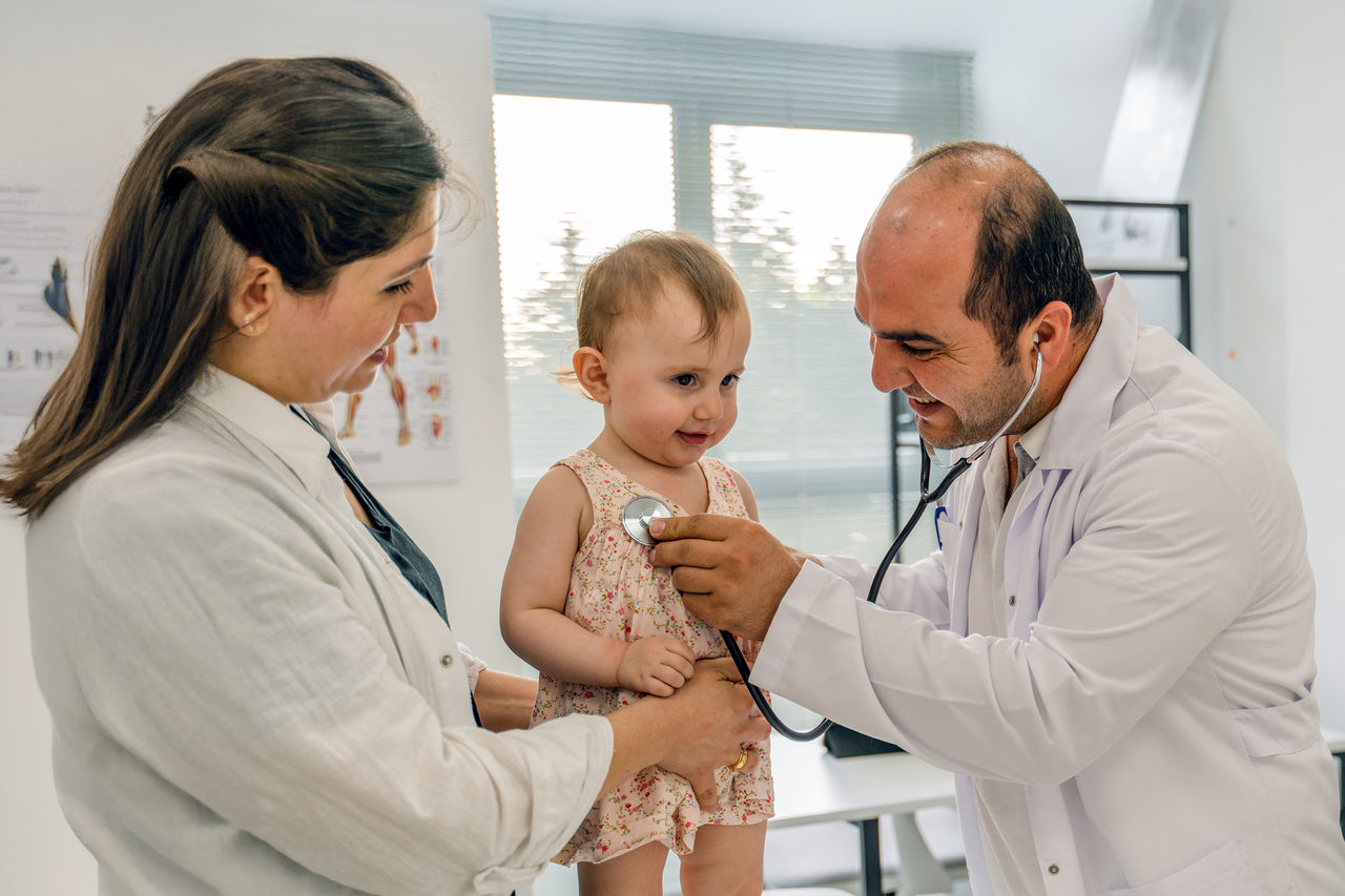 A healthcare provider uses a stethoscope to examine a young child standing on an exam table while another healthcare provider supports the child. The exam takes place in a bright medical office with windows and clinical equipment visible in the background.