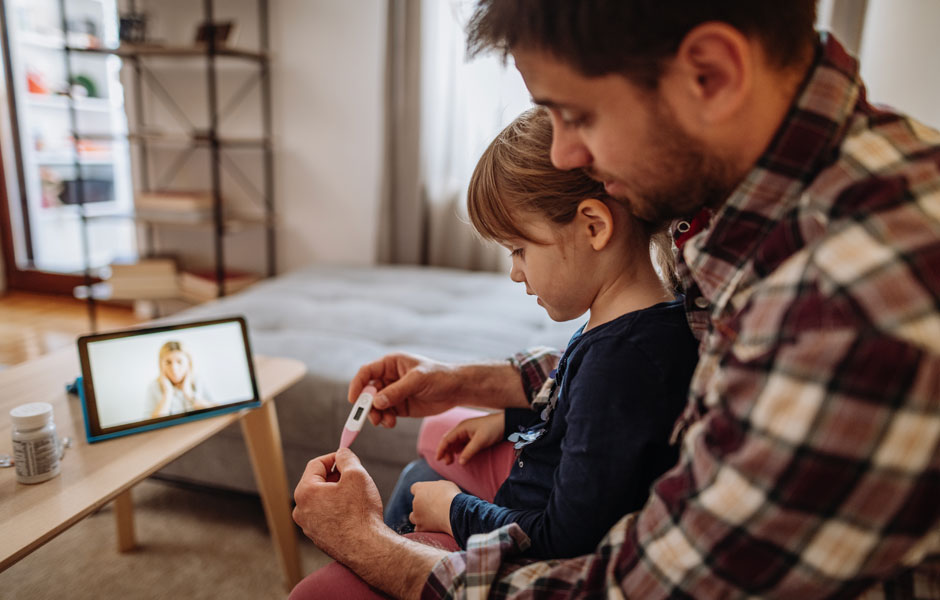 Dad holds young daughter on lap and checks thermometer while doctor in lab coat is on the smartphone screen in front of them for telehealth. 