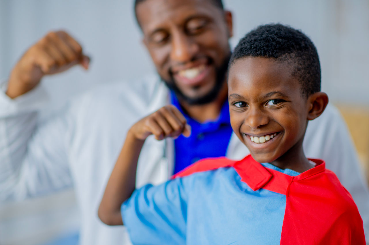 Boy wearing a cape flexing his bicep with a male doctor in the same pose behind him