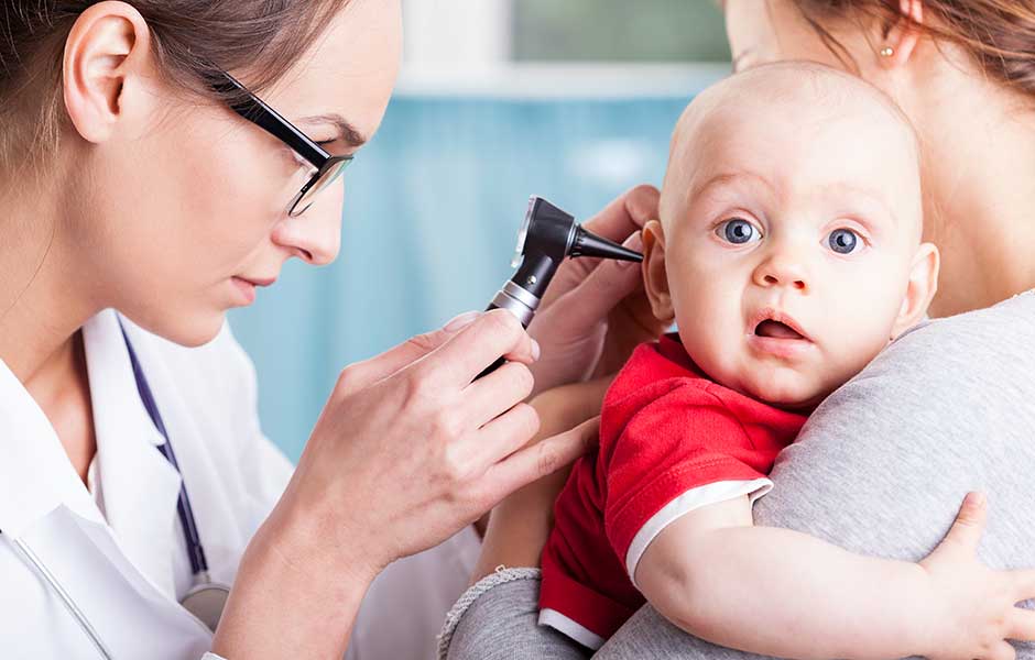 A doctor in a lab coat look into a baby's ear while mom holds the baby on her shoulder. 