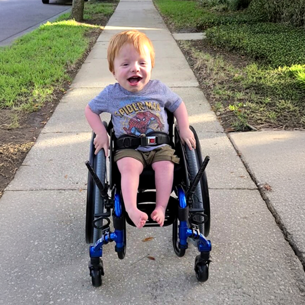 Young boy wearing a Spider-Man shirt smiling in a wheelchair on a sidewalk